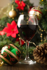 Glass of red wine on wooden table with Christmas decorations. Christmas tree and lights in the background.