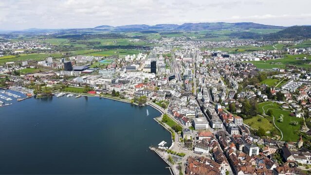 panoramic cinematic view of beautiful swiss lakeside city surrounded by lush green landscape and alpine farmland in the Swiss Alps featuring idyllic Swiss Lake of Zug with glassy crystalline water