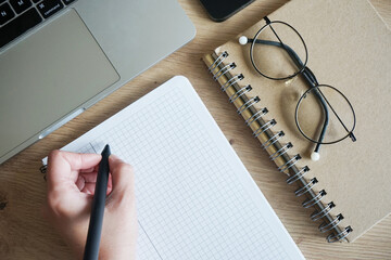 A man writes with his left hand in a notebook, and next to it lies a notebook, glasses, an open laptop and a smartphone