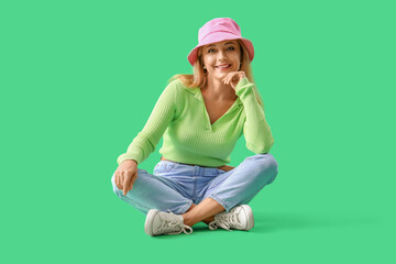 Stylish mature woman in bucket hat sitting on green background - obrazy, fototapety, plakaty