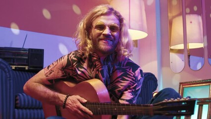 Horizontal shot. Hipster man sitting on floor. Extravagant male playing guitar at disco styled room. Man wearing sunglasses and smiling at camera. Long haired man relaxing at retro party.