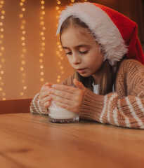 Cute little girl in Santa hat drinks milk from a glass at home. Xmas home holiday celebration