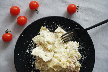 Pasta carbonara with parmesan cheese on a black plate with a black fork next to cherry tomatoes on a gray tablecloth