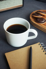 White cup with coffee next to cookies on a wooden tray, a notepad and a music column on a gray table