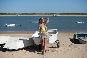 Obraz premium Pretty young blonde woman sitting in the fishermen's boats on the seashore. In the background on the horizon the blue sea and the boats working in the sea. Holiday and travel concept.