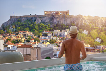 A tourist man in a swimming pool enjoys the view over the old town of Athens, Greece, and the Acropolis during his summer vacations