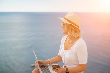Fototapeta premium Freelance women sea working on the computer. Good looking middle aged woman typing on a laptop keyboard outdoors with a beautiful sea view. The concept of remote work.