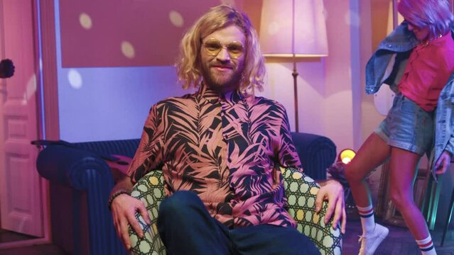 Horizontal shot. Hipster man twirling around sitting on chair. Hippy retro party. People attending disco party. Pink haired female dancing at background. Young people hanging at retro party.