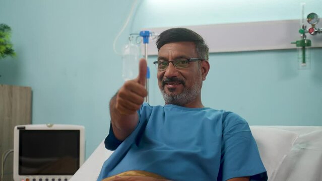 Indian Elderly recovered Patient showing thumbs up while sleeping on hospital bed after surgery by looking at camera - concept of successful diagnosis, approval and medical treatment.