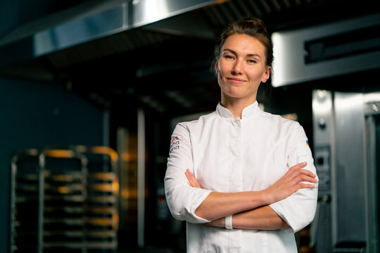 Portrait of a smiling female chef baker in a professional uniform in a bakery against background of an oven and fresh baked goods