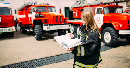 Female firefighter in in a protective suit with documents standing ain the background of a fire engines. Fire station