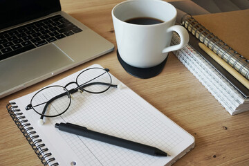 Notepad with pen and glasses, folders, open laptop and coffee cup nearby on wooden table