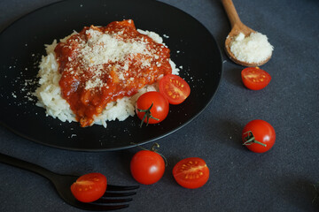 Rice with tomato sauce, meatballs and parmesan on a black plate next to a fork and cherry tomatoes on a gray tablecloth