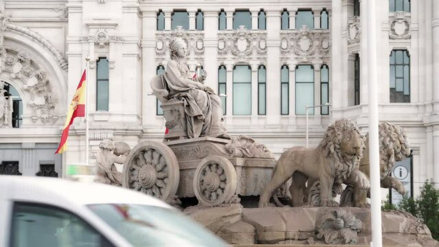 Timelapse at statue in the Plaza de Cibeles with the representation of the goddess of the same name and two lions with Spanish flags waving in the wind behind and traffic passing by
