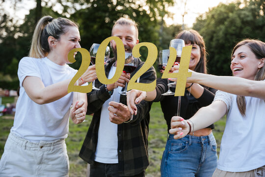 Group of friends at a party celebrate happy new 2024 year with elegant signs gold text - People have fun together outdoors toasting red wine glasses and smiling - Copy space