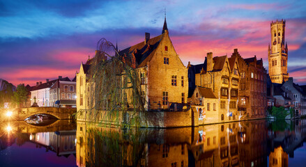 Fototapeta na wymiar Bruges, Belgium. Evening sunset with blue sky. Water channels of ancient medieval town view to Belfort van Brugge tower, famous landmark.