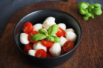 Mozzarella with cherry tomatoes in a black plate with basil on a wooden board on a dark gray background