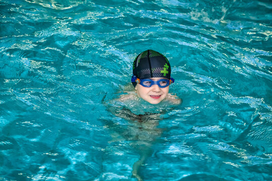 Kid boy swimmer in swimming goggles and swim hat in water pool workout, pensive looking. Child 5 year old in sport exercising in swim pool, learn. Sports children training concept. Copy ad text space