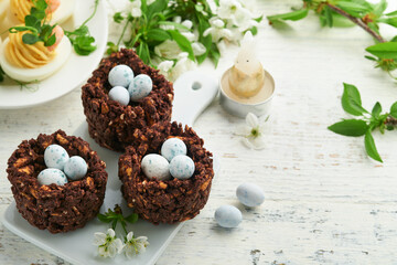 Obraz na płótnie Canvas Easter brunch or breakfast. Easter chocolate nest cake with chocolate candy eggs, traditional hot cross buns and deviled eggs with bouquets of blooming apple trees. Spring Easter holiday food concept.