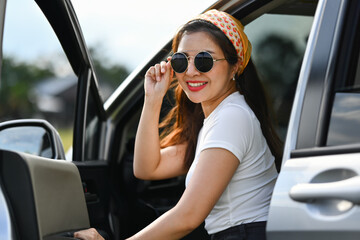 Confident female traveler wearing sunglasses sitting in car with open door. Travel and vacation concept