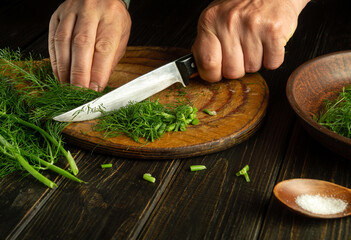 Slicing fennel or dill with a knife in the hands of a chef on a kitchen cutting board before...