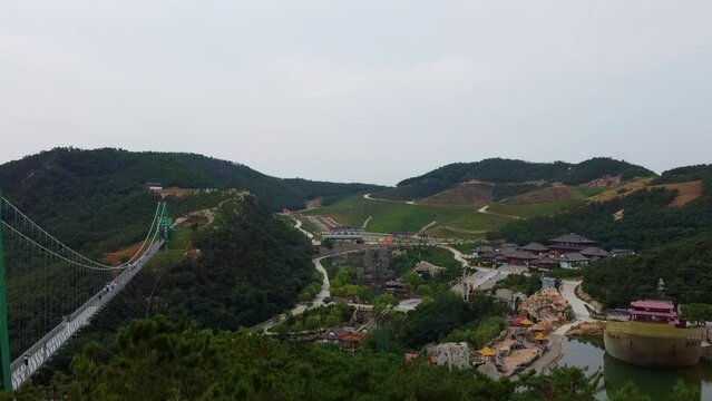 Aerial ascending view behind trees revealing suspension glass bridge and beautiful scenery at Huaxiacheng theme park in Weihai, China