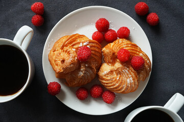 Cakes with cream and raspberries on a white plate next to a cup of coffee and a knife on a dark background