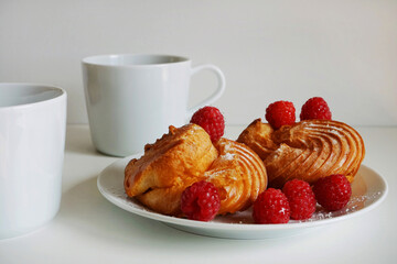 Cakes with cream and raspberries on a plate next to a cup of coffee and a knife on a white background