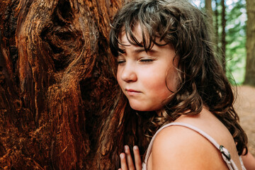 Calm little girl touching old tree trunk in forest