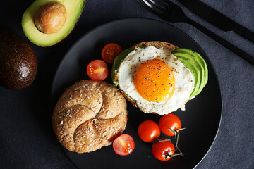 Fried eggs, avocado, cherry tomatoes with bread on a black plate on a dark background