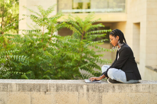 Focused ethnic businesswoman working on laptop while sitting at stone border near plants