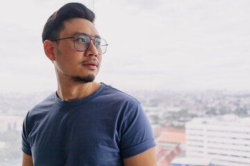 Asian man wear glasses and blue t-shirt with beard, smiling and standing over city view at condominium, looking out side at apartment room alone, having happy good life.