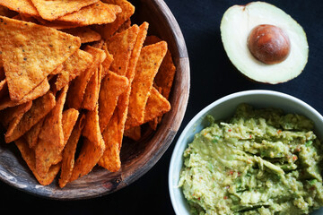 Plates with nachos and guacamole next to avocado on black background