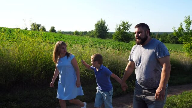 Young parents with little son hold hands of each other walking along rural road near green meadows. Mother and father with child going along path near cultivated fields. Family spending time together