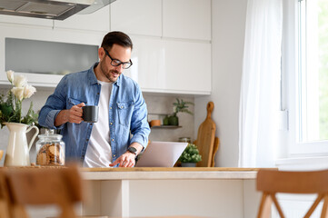 Focused businessman holding coffee cup and reading e-mails over laptop on kitchen counter at home