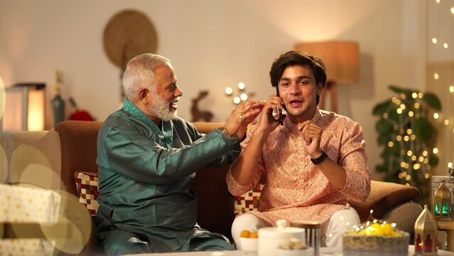 Indian father and son talking on phone on diwali.they both are wearing traditional dress.father happily snatching the phone from his son to talk while both are sitting on the sofa.