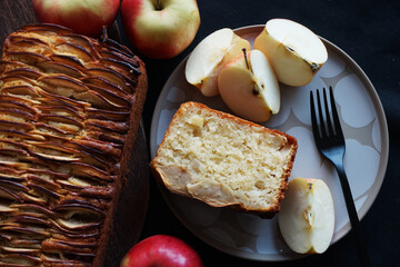 Apple pie next to a piece of pie on a plate and apples on a dark background