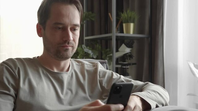 The man looks indifferently at the smartphone screen, scrolling through the contents while at home. Signs of dopamine addiction from constant viewing of entertainment content on social networks.