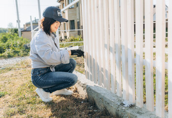 beautiful asian woman paints a wooden fence with white paint.