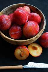 Ripe plums in a bowl next to sliced ​​plums and a knife on a dark tablecloth