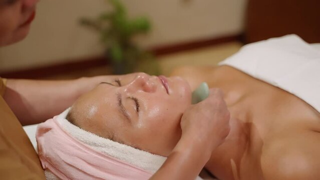 Young caucasian woman gets lifting facial massage with a gua-sha tool at spa and wellness center. Female client experiences facial lymphatic drainage therapy using jade stone and massage oil closeup