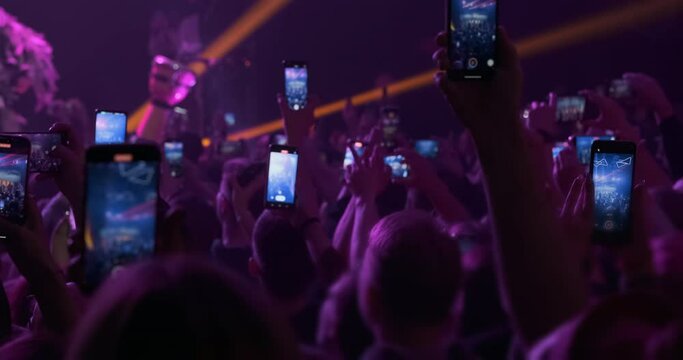 Fans film by phones band concert on pink light background. Blue phone screens in crowd people hands illuminated by pink magenta, orange spotlight. Audience dance in music festival. Entertainment event