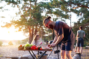 A company of young people prepares a vegetable salad in nature against the backdrop of an evening...