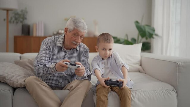 Old senior grandpa cute little child grandson gamers team hold controllers playing video game sit on sofa at home. Family two generations play gamepads looking at screen. Leisure, pastime concept.