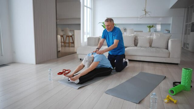 Senior couple man woman training at home together. Wife sitting on mat doing stretching exercises bending to knees, husband helping her press on back. Sport fitness active healthy lifestyle concept.