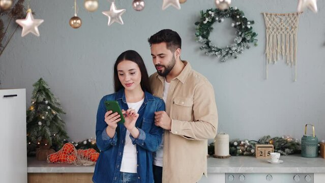 Couple in love hold mobile phone enjoy winter Christmas New Year holidays at modern cozy ornate kitchen. Modern tech usage, spend free time online, fun on internet, use new app on xmas Eve concept.
