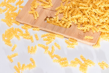 Obraz premium Uncooked Fusilli Pasta Scattered on Wooden Board and White Table. Raw and Dry Macaroni. Fat and Unhealthy Food