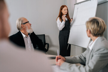 Professional female coach gives corporate presentation for businesspeople in office. Red-haired...