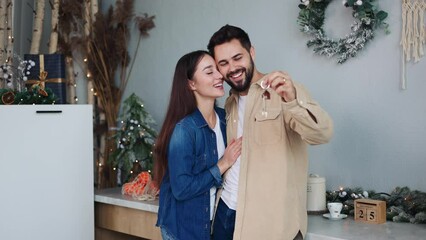 Loving couple in kitchen by Christmas tree, proudly displaying keys to their new apartment. Newlyweds celebrate buying their first home, special New Year gift. Symbol of new beginnings in real estate