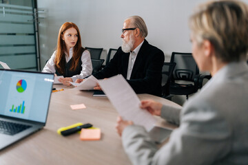 View from shoulder of unrecognizable middle aged female mentor, corporate teacher teaching employees to analyze project documents, reviewing reports. Employees brainstorming at conference.